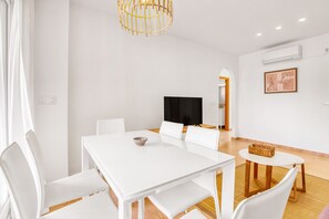 A modern, well-lit room with a white dining table, white chairs, a television, and a small round table with a basket.