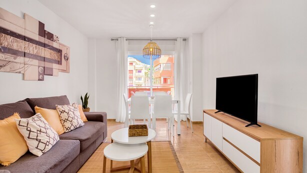 En moderne stue med sofa, tv på et træstel, spiseplads med stole og udsigt til bygninger.
