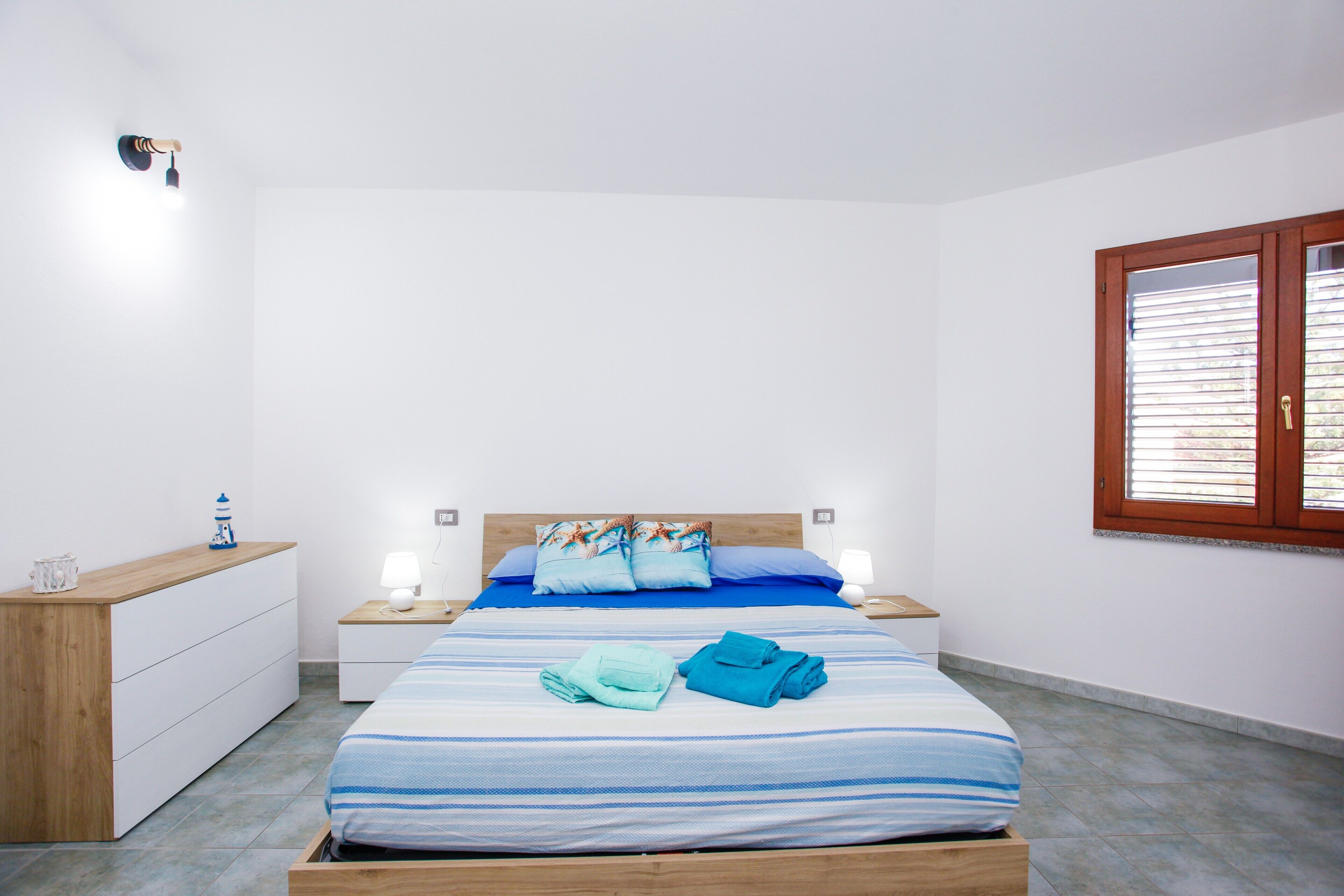 A bedroom with a bed, two bedside tables, a nightstand, a wooden dresser, and a window with blinds.
