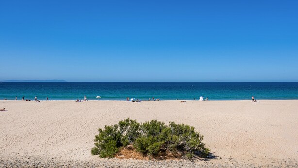 A beach with people sunbathing and swimming, clear blue sky, and calm sea.