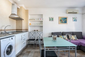 A compact living space with a washing machine, a dining table, a sofa, and a small kitchen area with a sink and cabinets.