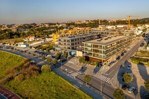 Un immeuble à plusieurs étages, doté d’une architecture moderne, comprenant des logements et des espaces commerciaux, un parking et une route où circulent des véhicules.