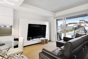 A modern living room with a flat-screen TV, a glass dining table, a sofa, and a large window offering a view of buildings.