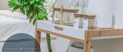 A neatly organized room with a bed, a bedside plant, a wooden shelf with kitchen items, and a wall-mounted lamp.