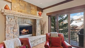 Une cheminée en pierre avec un feu de bois, deux fauteuils en cuir avec des coussins et une grande fenêtre donnant sur une terrasse.