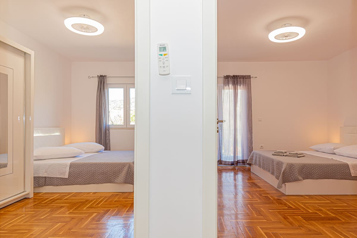 A hotel room with two beds, a wooden floor, a window with curtains, and a thermostat on the wall.