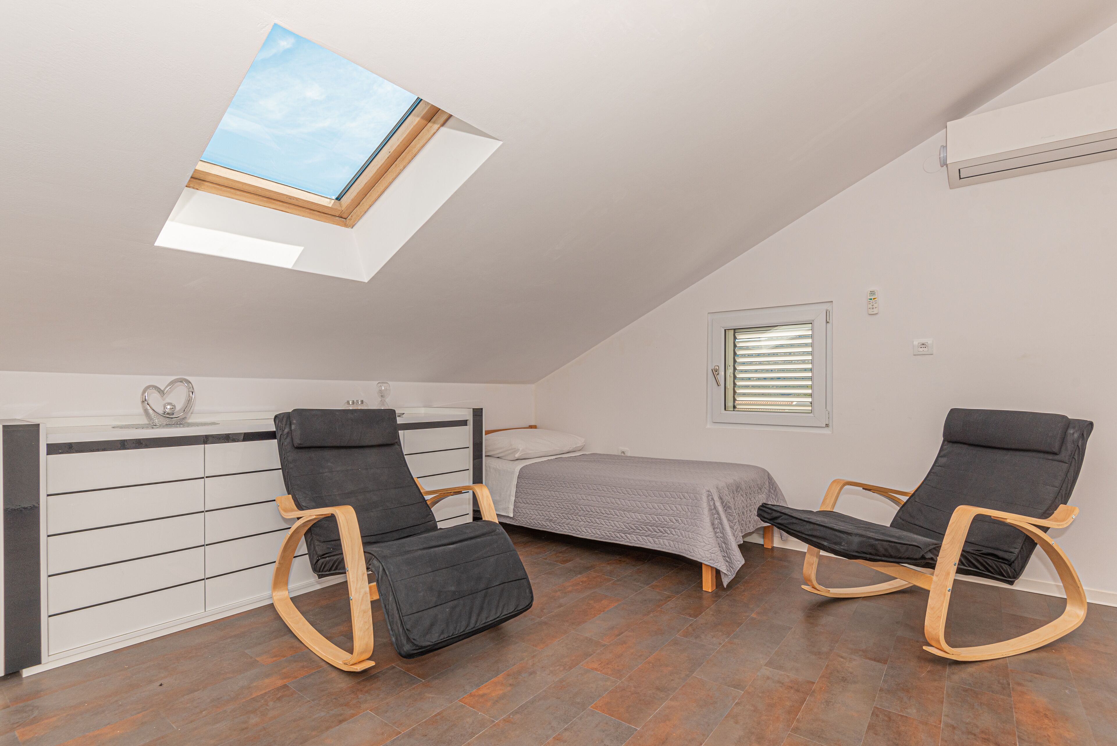 Een compacte kamer met dakraam, een bed, twee schommelstoelen, een klein bureau en een raam met jaloezieën.