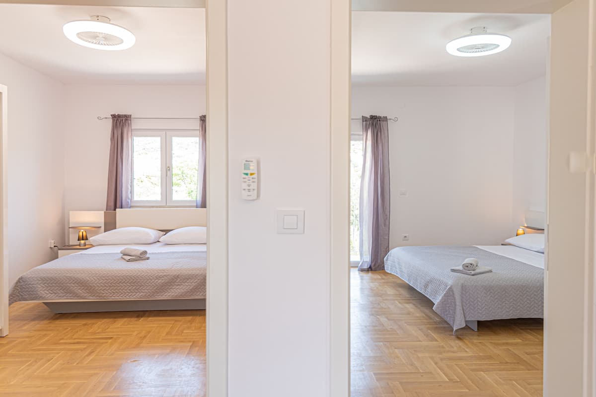 A bedroom with two beds, a bedside table with a lamp, a window with curtains, and a thermostat on the wall.
