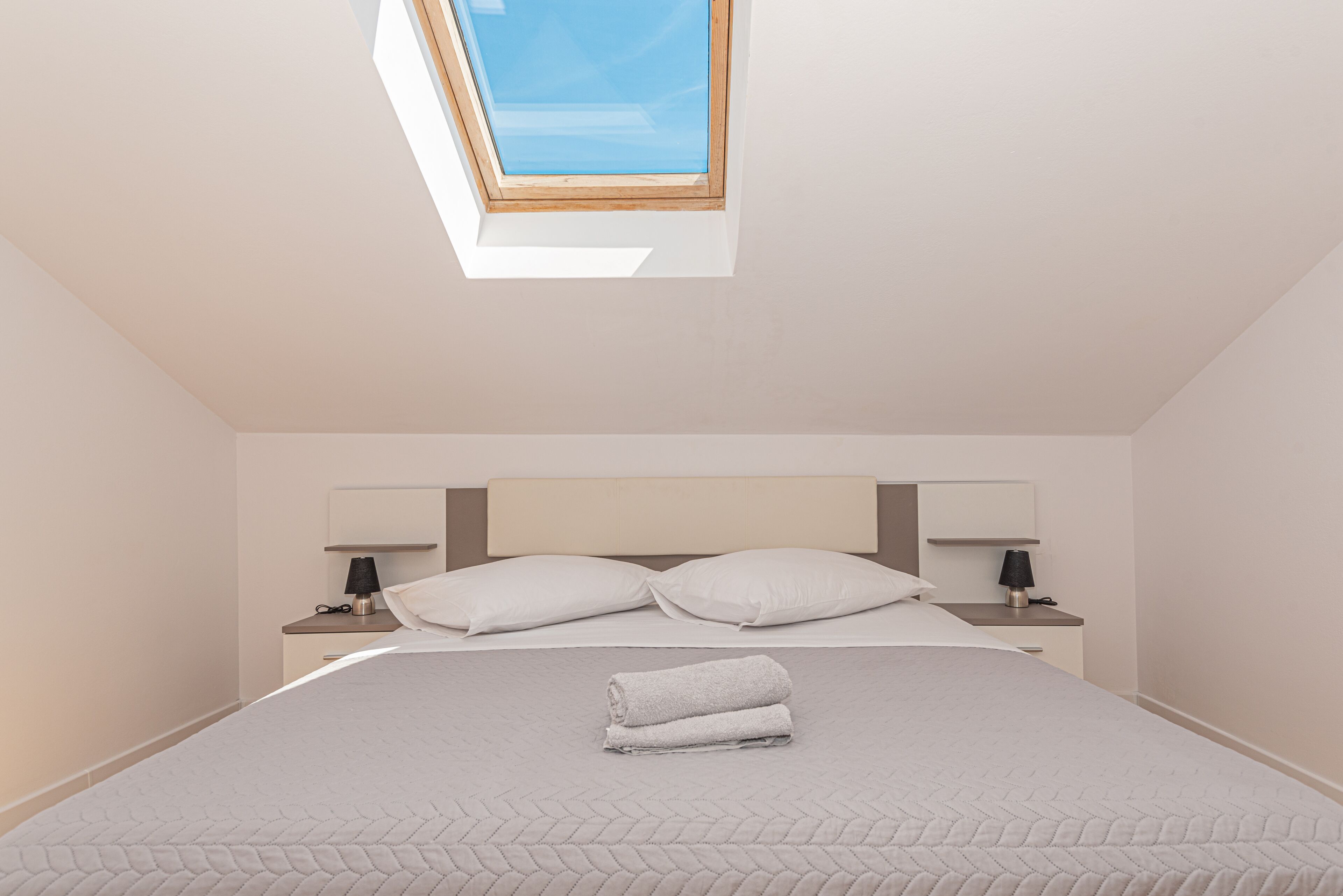 A bedroom with a skylight, a bed with a rolled towel, two bedside tables with lamps, and a headboard.