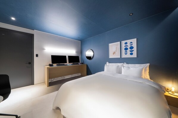 A modern hotel room with a large bed, a desk with a computer, and a wall-mounted TV.