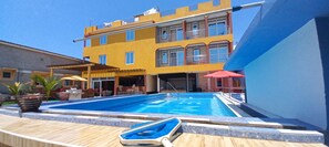 A pool area with a blue pool, wooden deck, and a yellow building with balconies. - Le Mach Apartments (Kilifi)