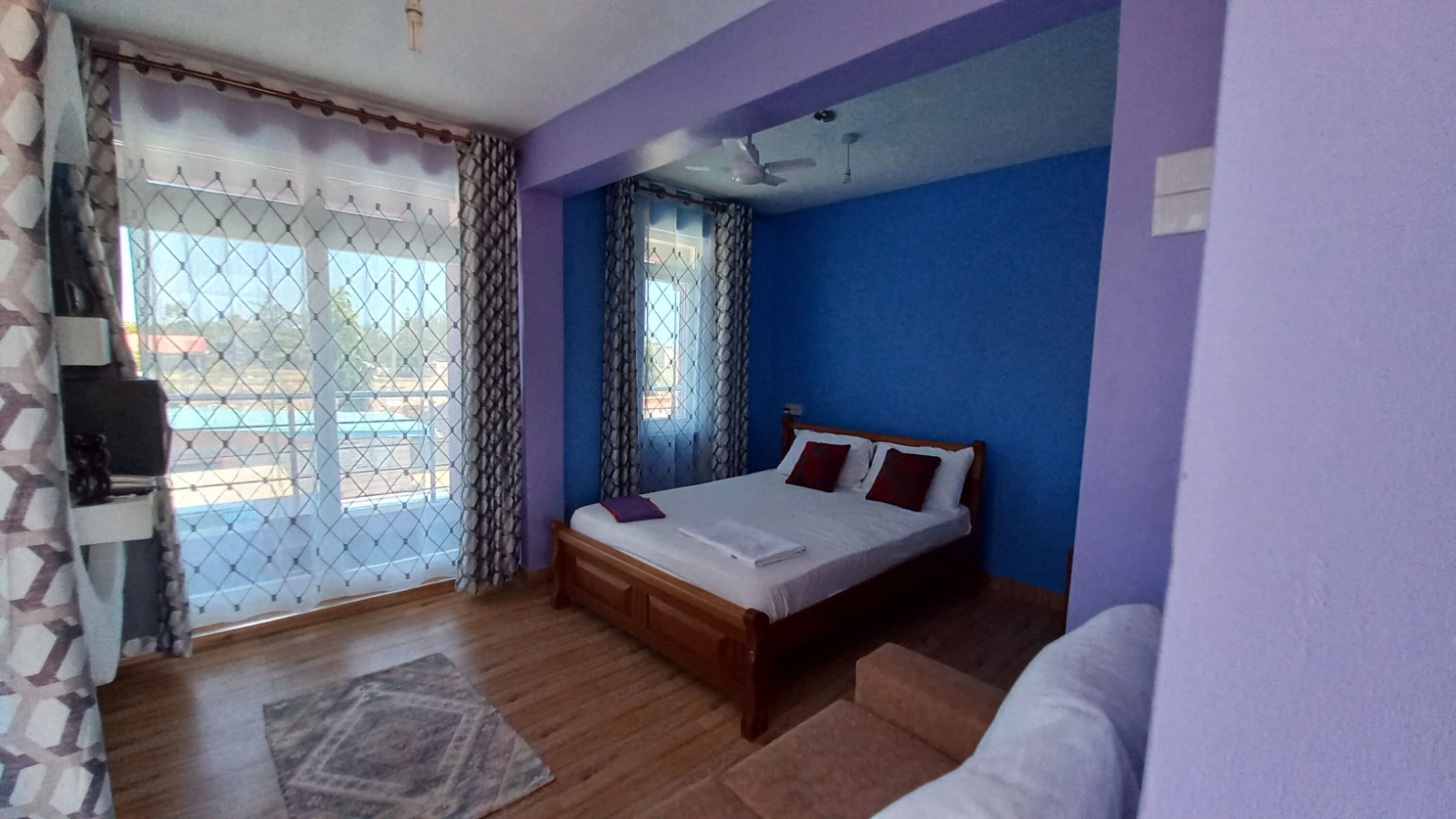 A bedroom with a wooden bed, a ceiling fan, and a view through a window with a chain-link fence.