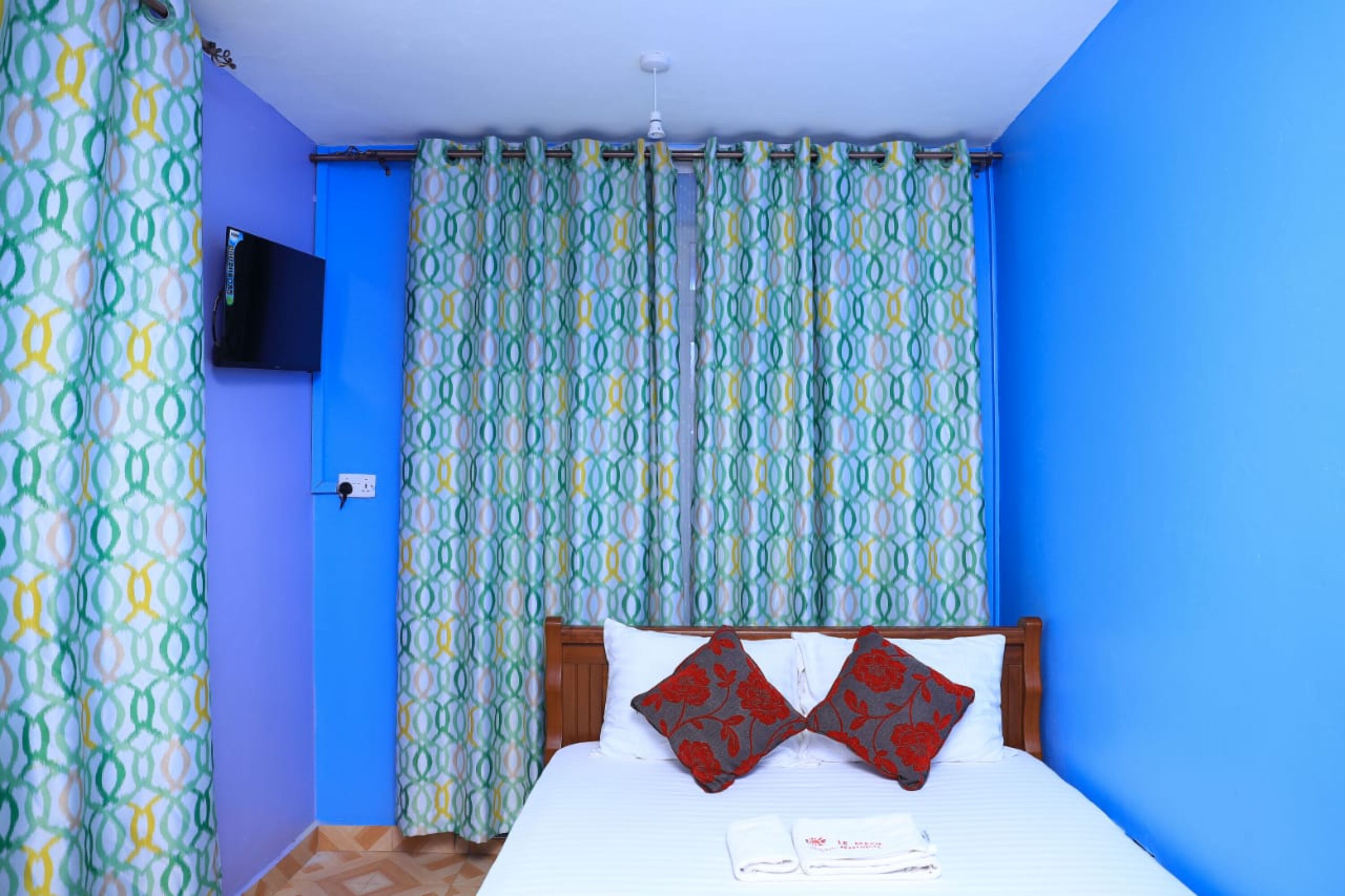 A room with a bed, patterned curtains, a mounted TV, and a blue wall.