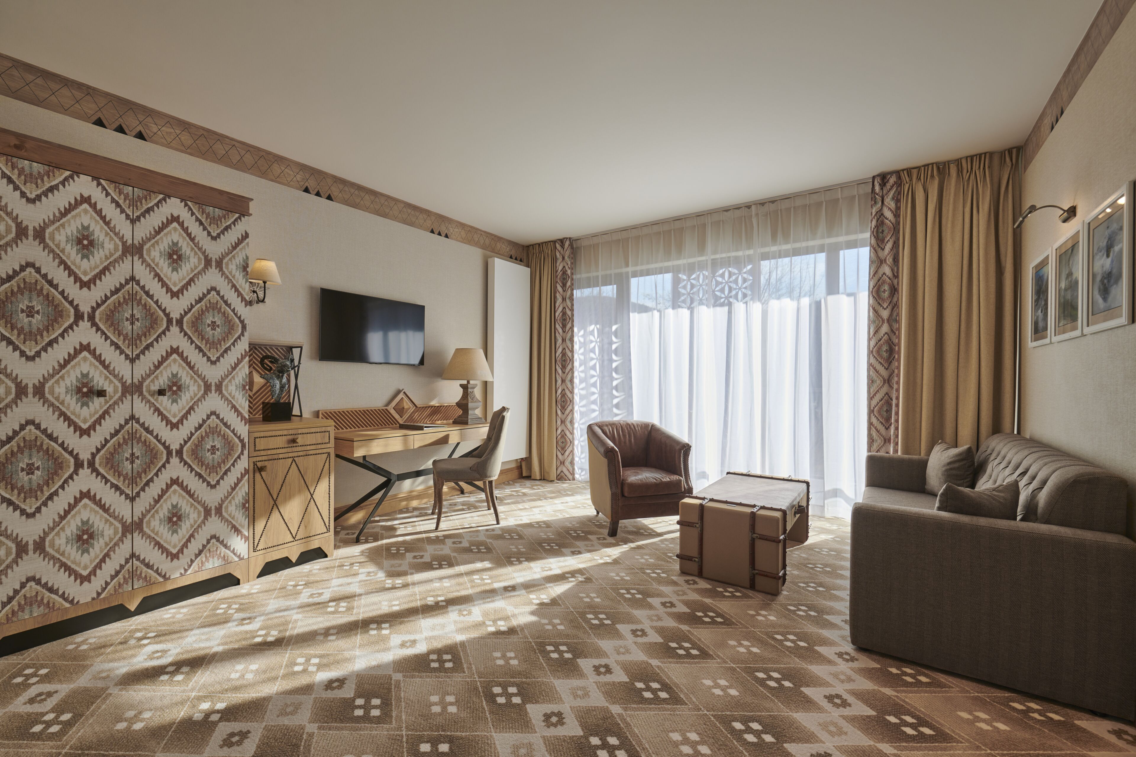 Una habitación de hotel con alfombra estampada, un sofá, una silla, un escritorio y un televisor en la pared.