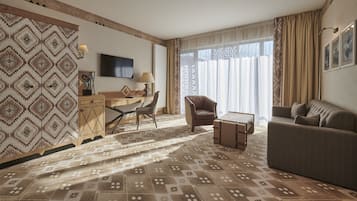 A hotel room with a patterned carpet, a sofa, a chair, a desk, and a television on the wall.