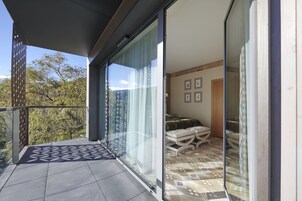 Uma varanda com piso estampado, uma porta de vidro deslizante que dá para um quarto com cama e tapete estampado, e vista para árvores lá fora.