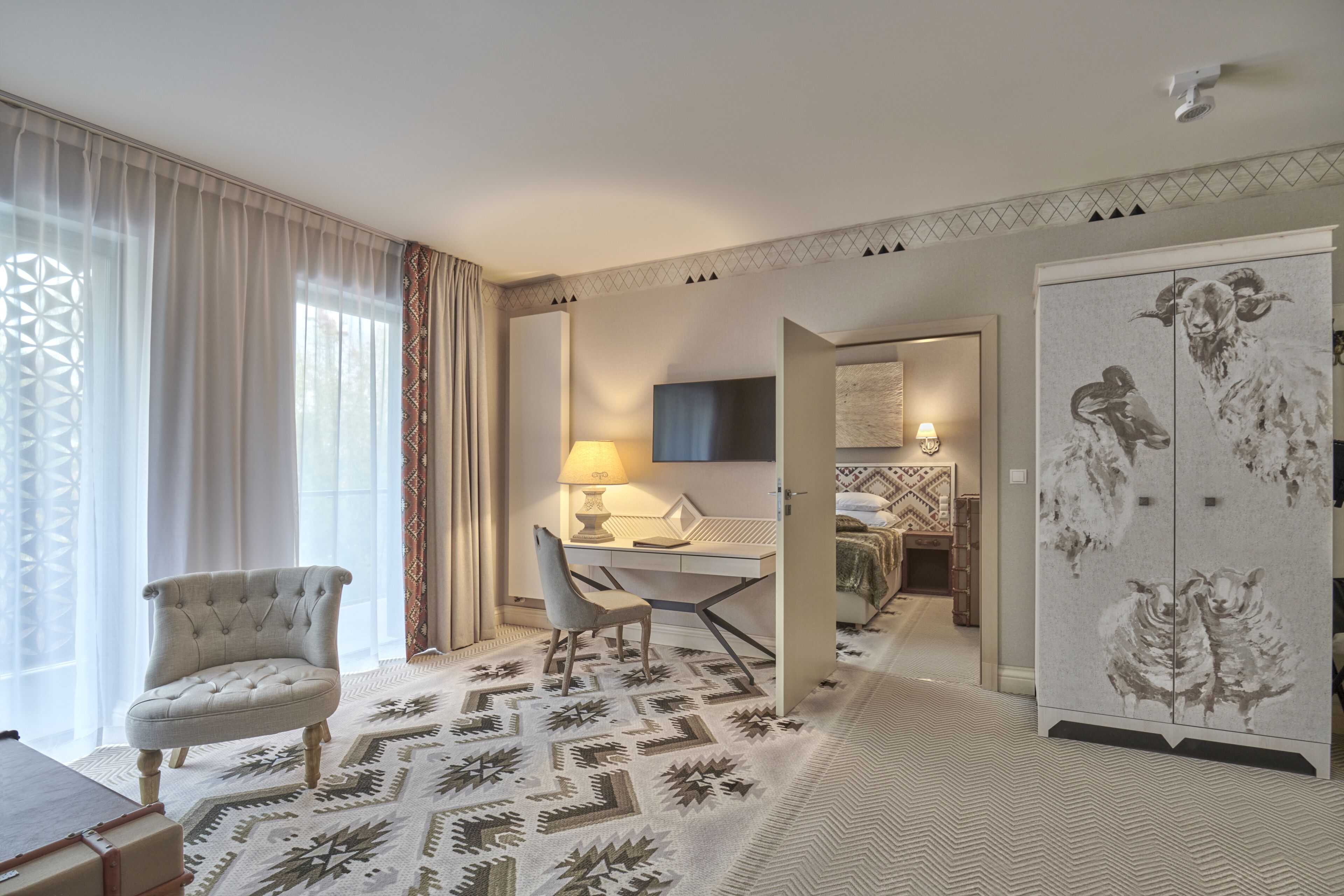 A hotel room with a tufted armchair, a desk with a lamp, a patterned rug, and a wardrobe with a sheep design.