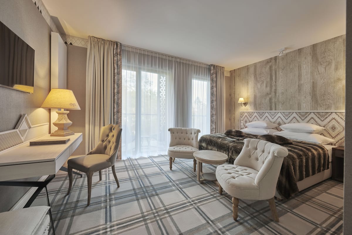 Habitación de hotel con cama, dos sillones, escritorio con lámpara y ventana grande con cortinas.