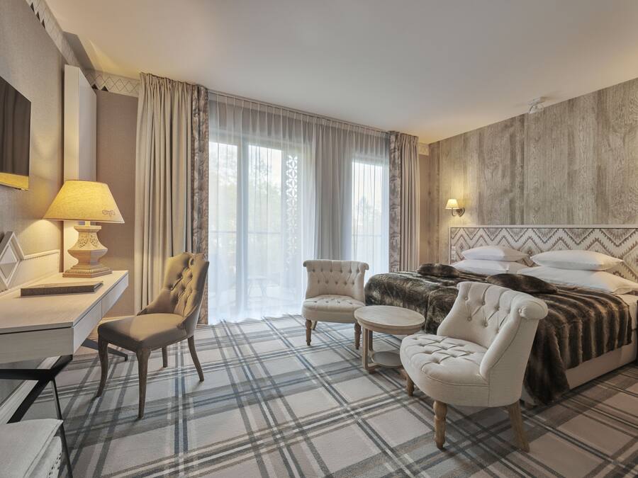 A hotel room with a bed, two armchairs, a desk with a lamp, and a large window with curtains.