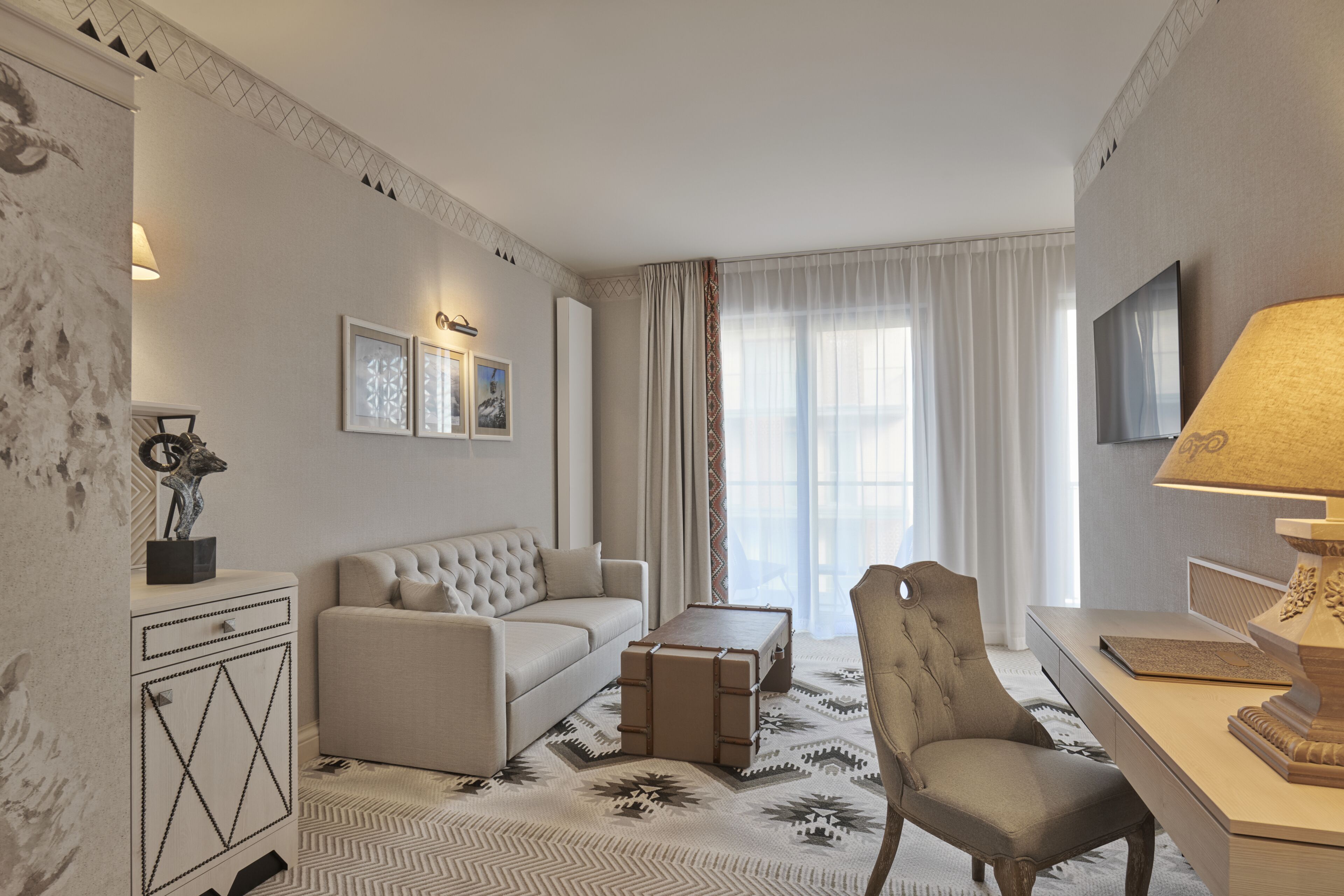 A modern hotel room with a sofa, armchair, and a desk with a lamp.