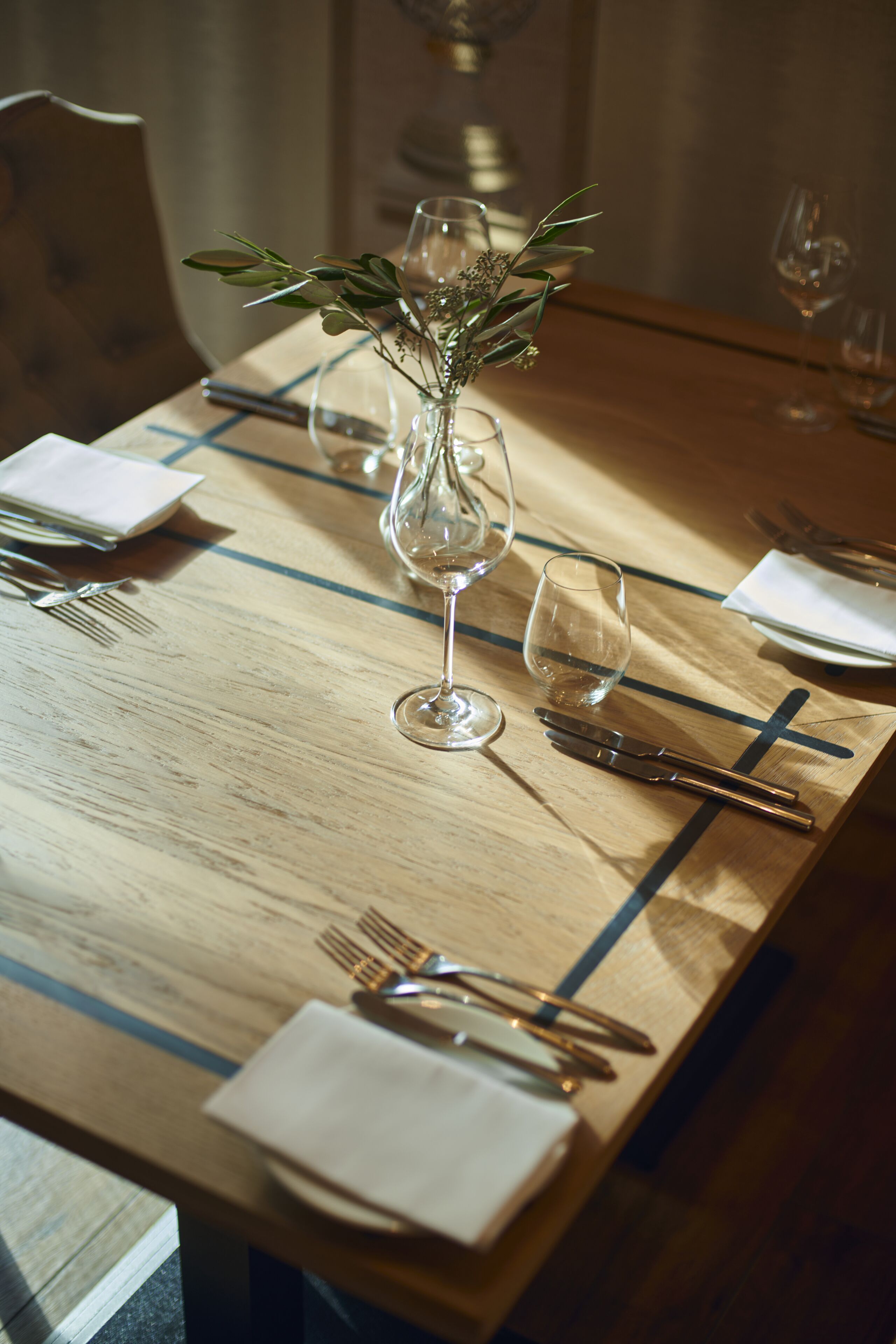 A wooden dining table with a vase of greenery, wine glasses, and cutlery.