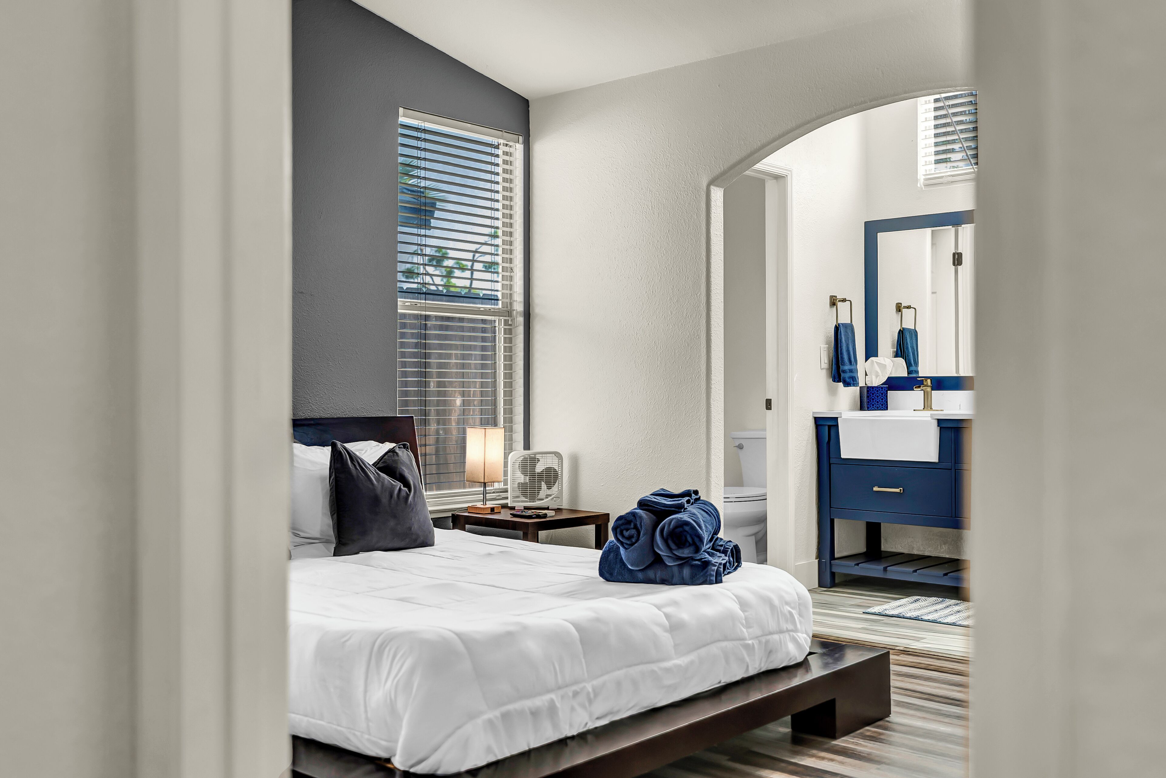 A modern hotel room with a bed, a bedside table with a lamp, a fan, and a neatly made bed with rolled towels.