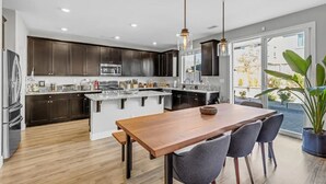 A modern kitchen with dark cabinetry, a central island, and a dining area with a wooden table and chairs. - New South Reno Home with Hot Tub (Reno)