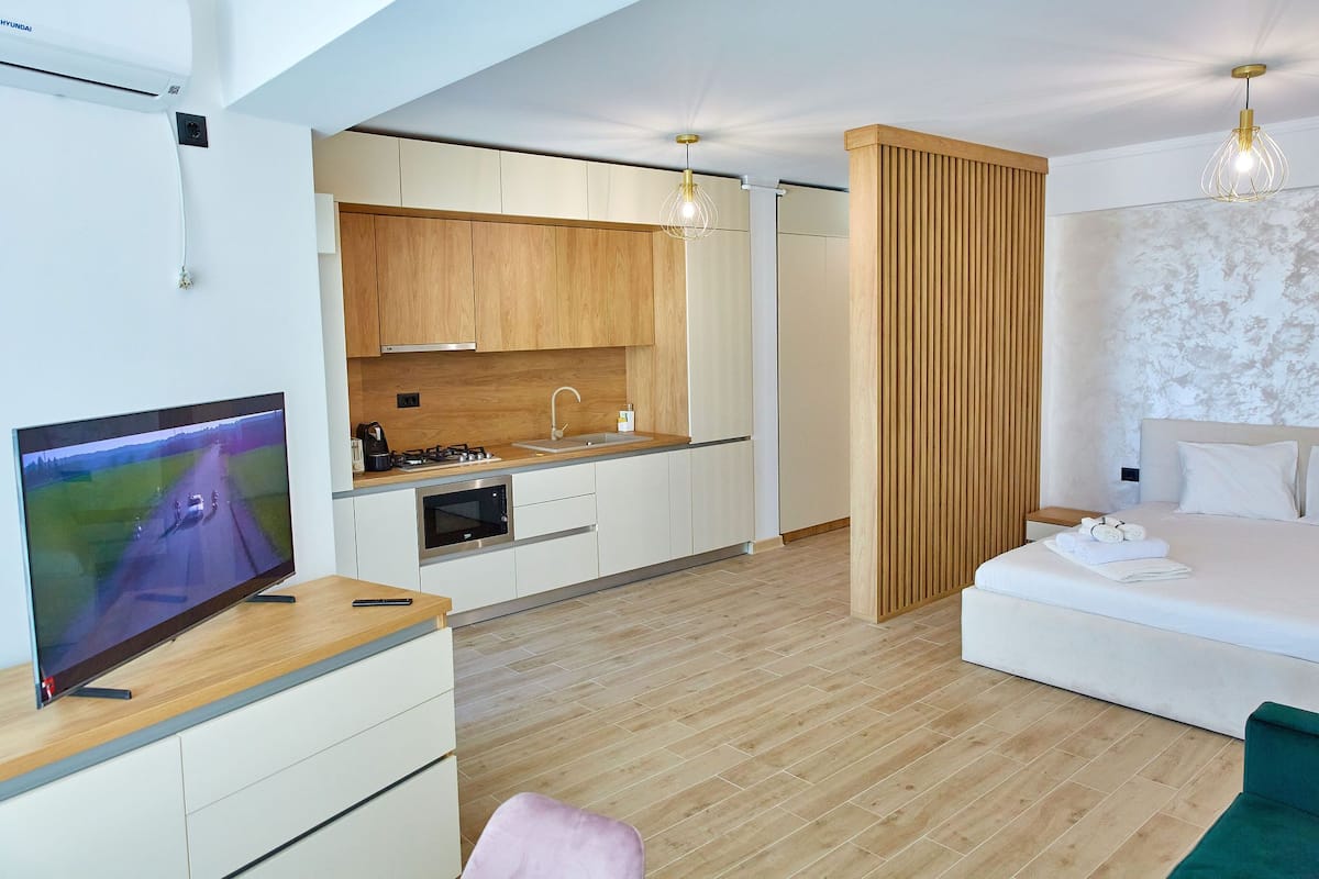 A modern hotel room with a wooden floor, a kitchen area with white cabinets, a flat-screen TV, and a bed with white linens.