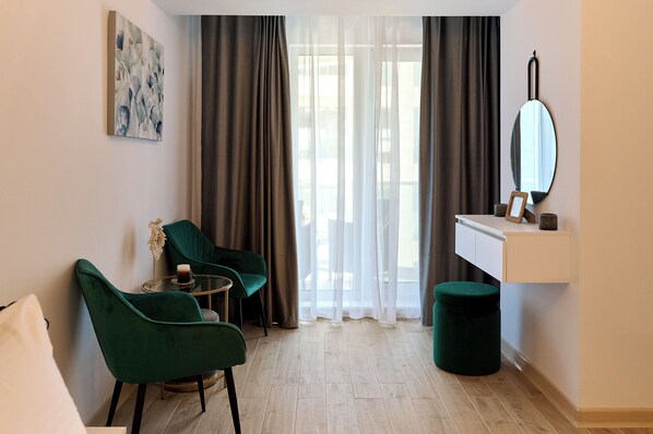 A modern hotel room with a green chair, a small round table, a mirror, and a green ottoman.