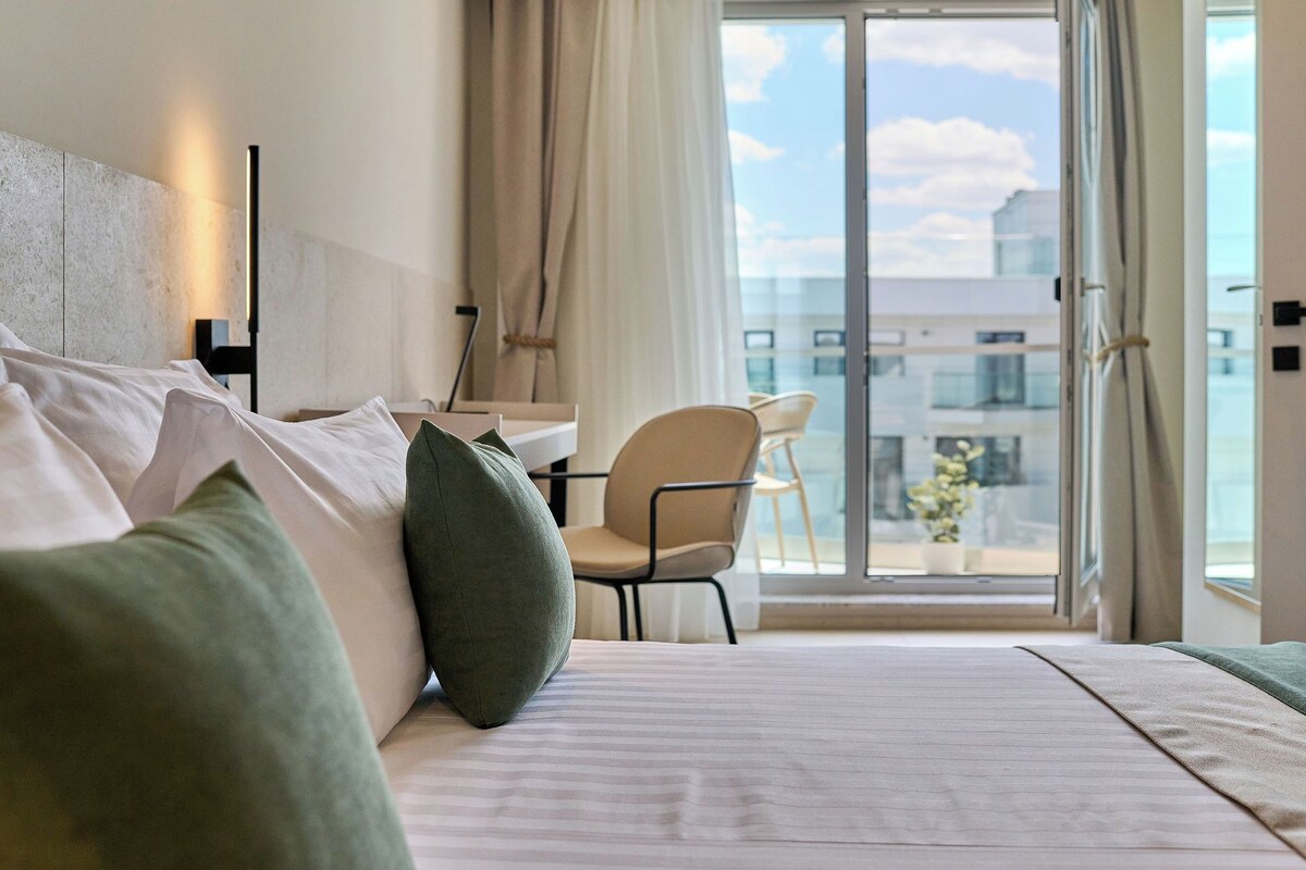 A hotel room with a bed, a desk, a chair, and a view of the city through a large window.