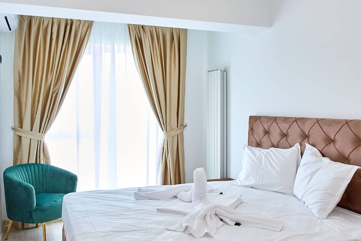 A hotel room with a large bed, beige curtains, a teal armchair, and a ceiling light fixture.