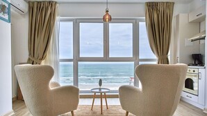 A room with two armchairs facing a large window overlooking the sea, a small round table, and a kitchen area to the right. - Alezzi Infinity Resort & Spa (Navodari)