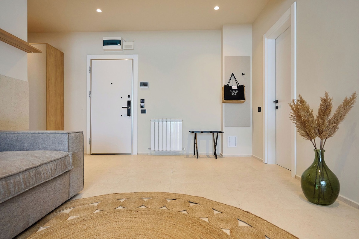 A modern hotel lobby with a grey sofa, a woven rug, a green vase with pampas grass, a radiator, and a small table.