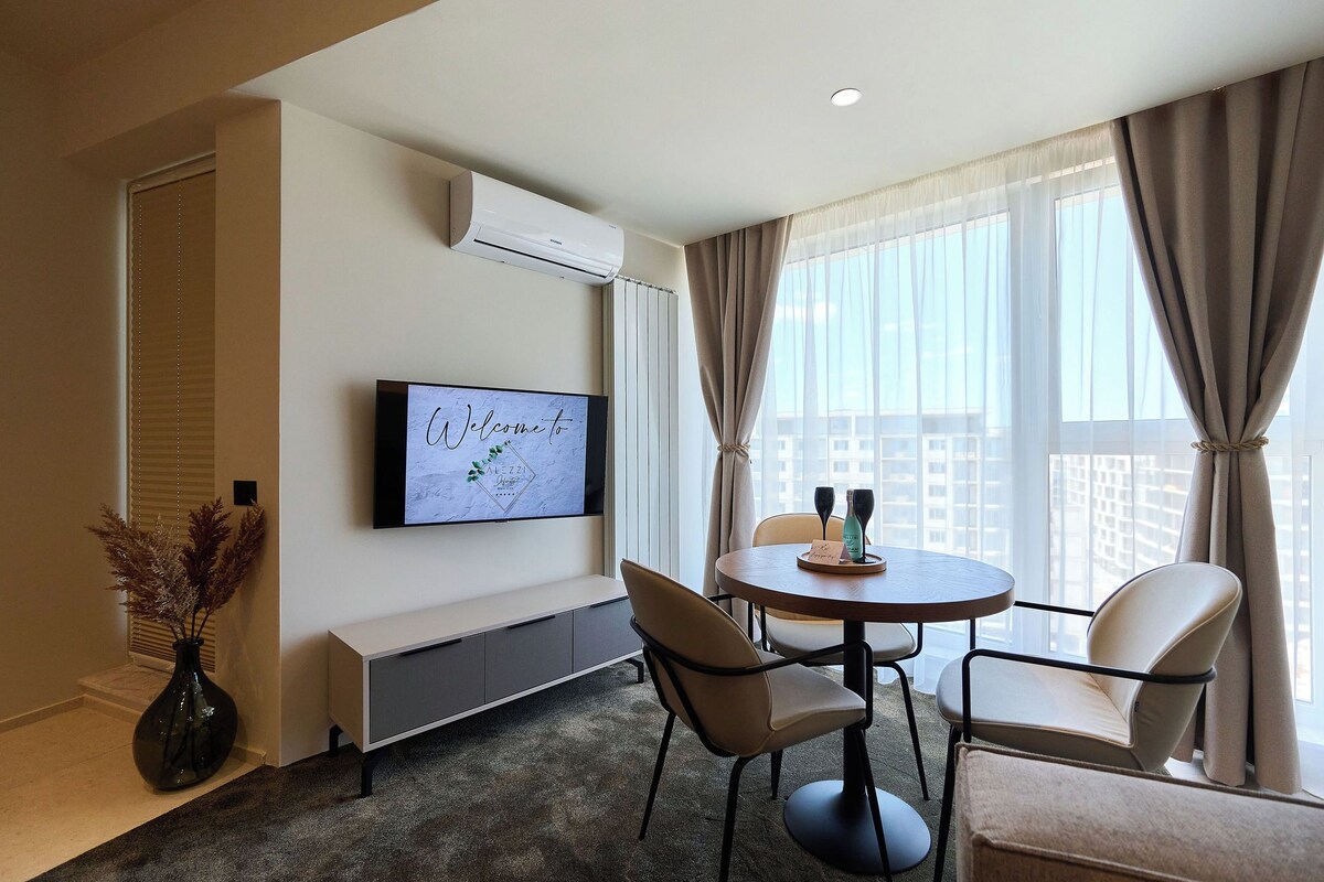 A modern hotel room with a flat-screen TV on the wall, a round dining table with chairs, a vase with dried pampas grass, and a large window with curtains.