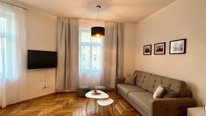 A modern living room with a herringbone wooden floor, a grey sofa, a white coffee table, and a TV mounted on the wall. - Firstclass 2r_apartment (Leipzig)