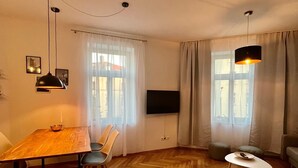 A modern living room with a wooden herringbone floor, a dining table with chairs, a TV mounted on the wall, and two pendant lights. - Firstclass 2r_apartment (Leipzig)