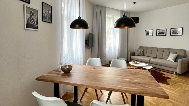 A modern dining area with a wooden table and white chairs, a sofa, and a dining table lamp. - Firstclass 2r_apartment (Leipzig)