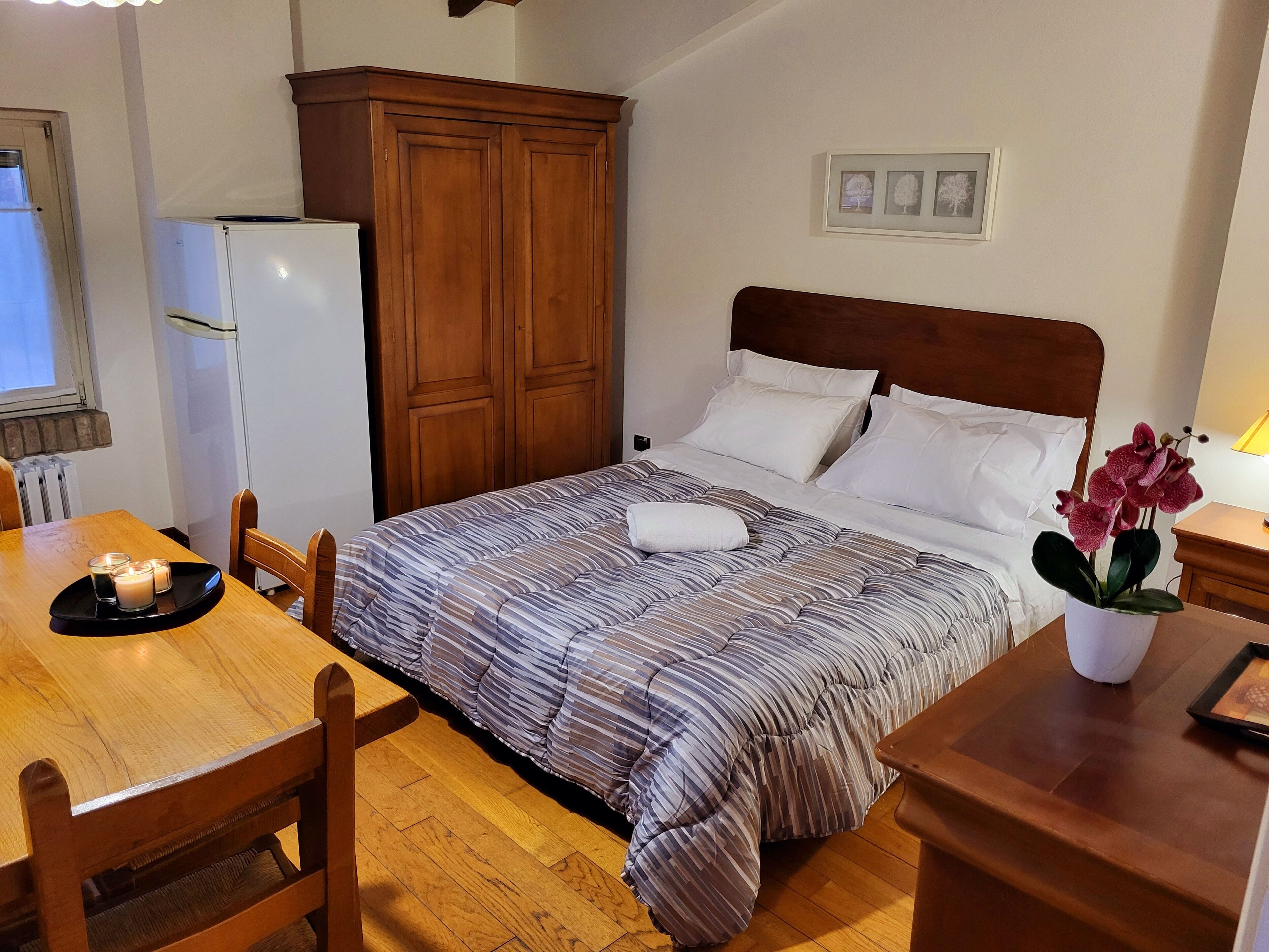 A bedroom with a bed, wooden wardrobe, a small table with candles, and a potted plant.