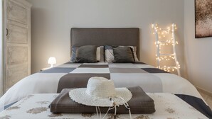 Una cama bien hecha con una colcha a cuadros, un sombrero blanco de ganchillo y una lámpara de noche.