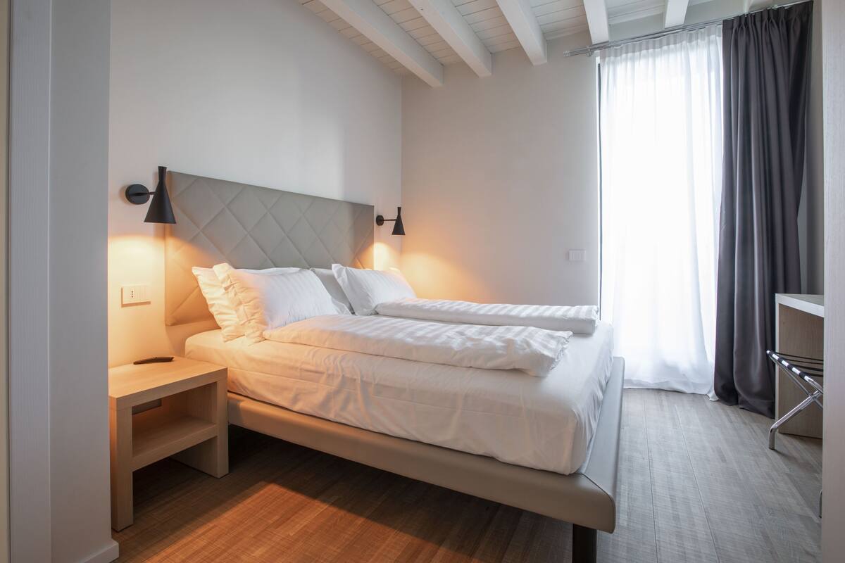Una cama doble con cabecera acolchada, mesitas de noche y una ventana con cortinas.
