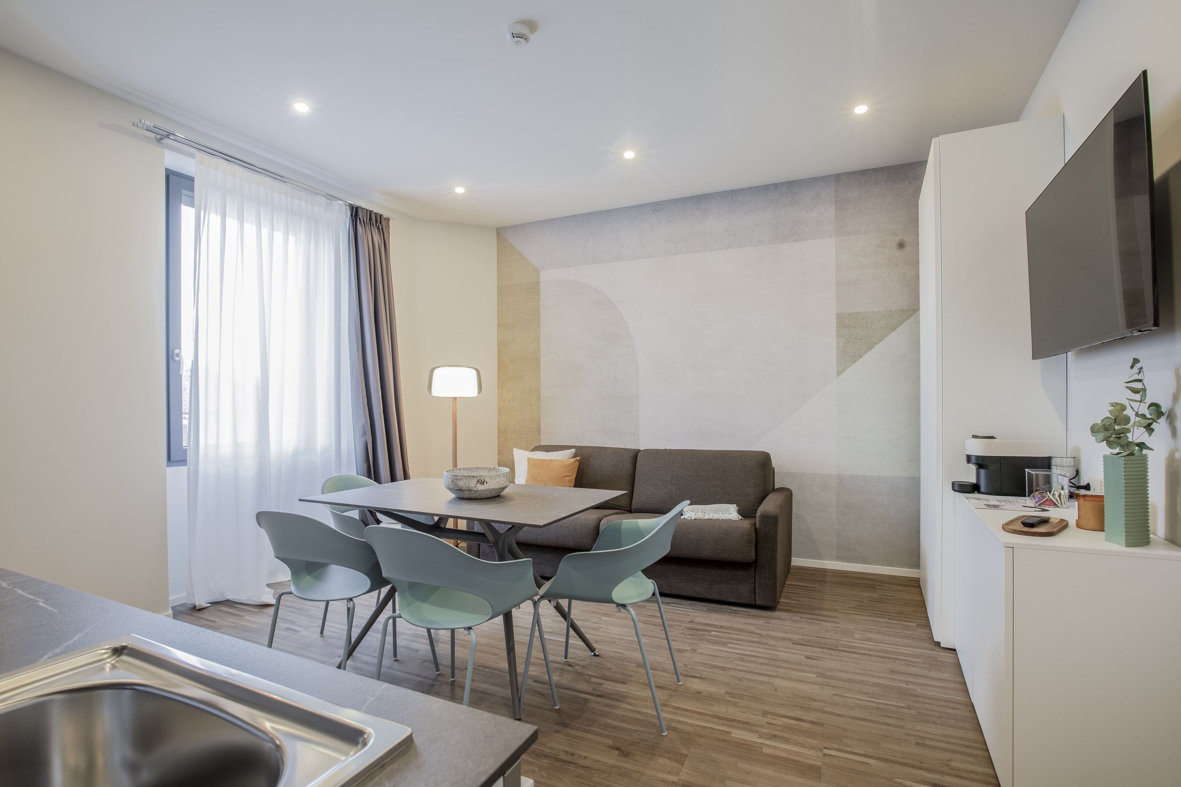 A modern kitchen with a sink, dining table, and chairs. There is a sofa and a flat-screen TV mounted on the wall.