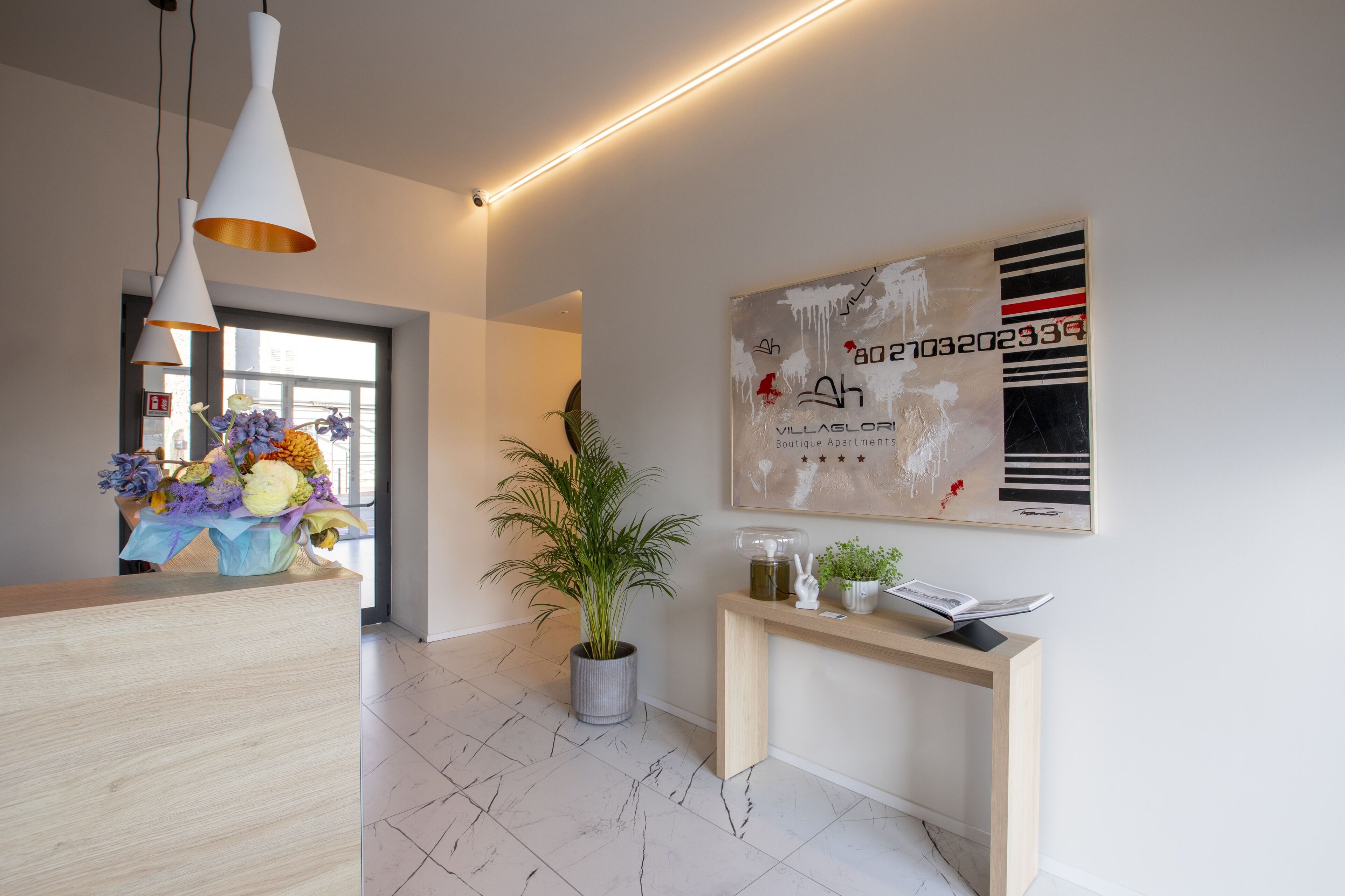 A modern reception area with a wooden reception desk, a potted plant, a console table with decorative items, and a large abstract painting on the wall.