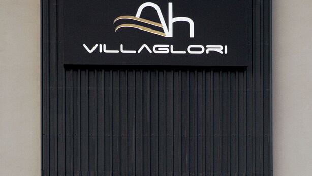 Mustalla pohjalla oleva kyltti, jossa lukee teksti ”VILLAGLORI”, ja tekstin yläpuolella on tyylitelty kirjain ’h’.