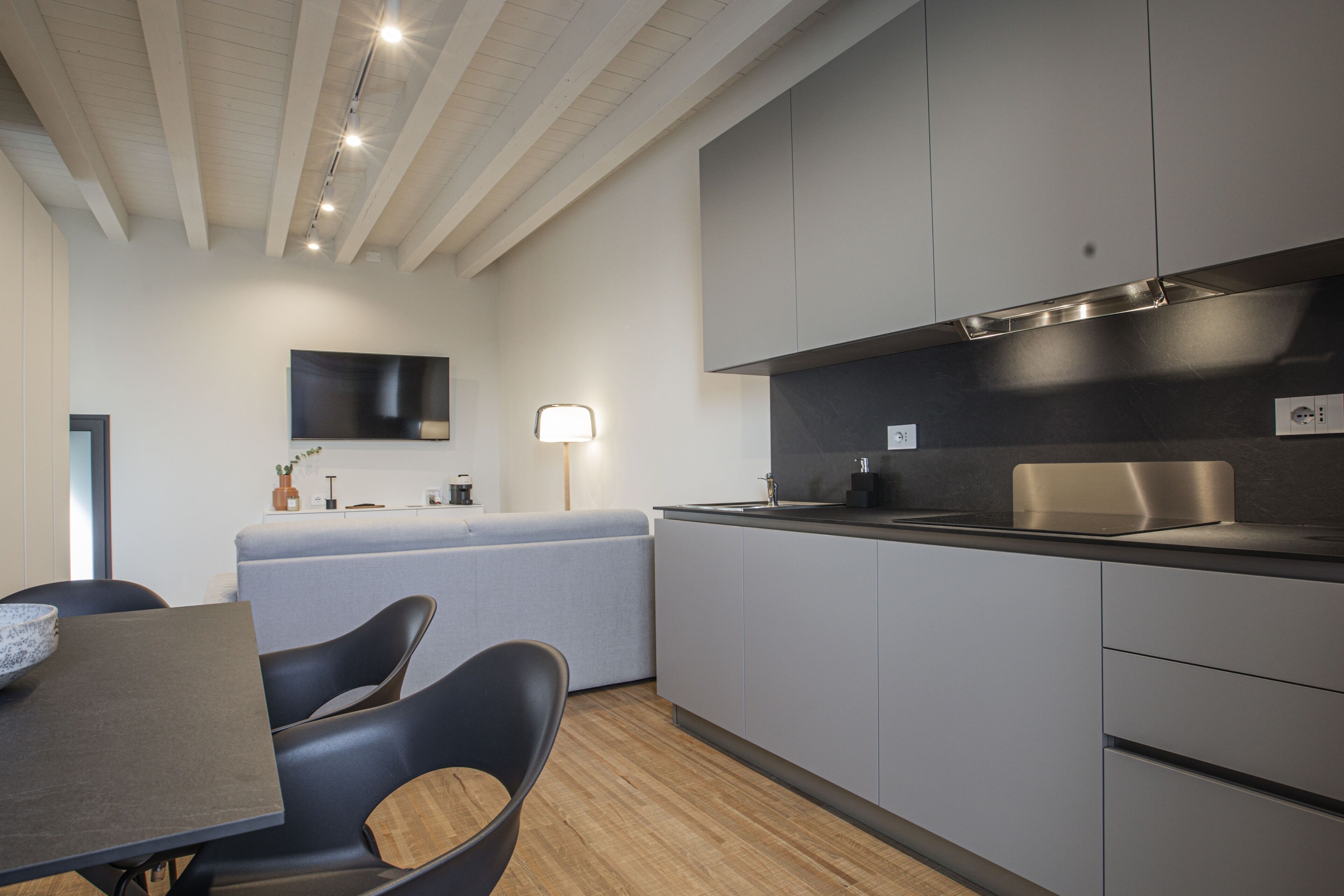 A modern kitchen with grey cabinets, a wooden dining table, and a flat-screen TV mounted on the wall.