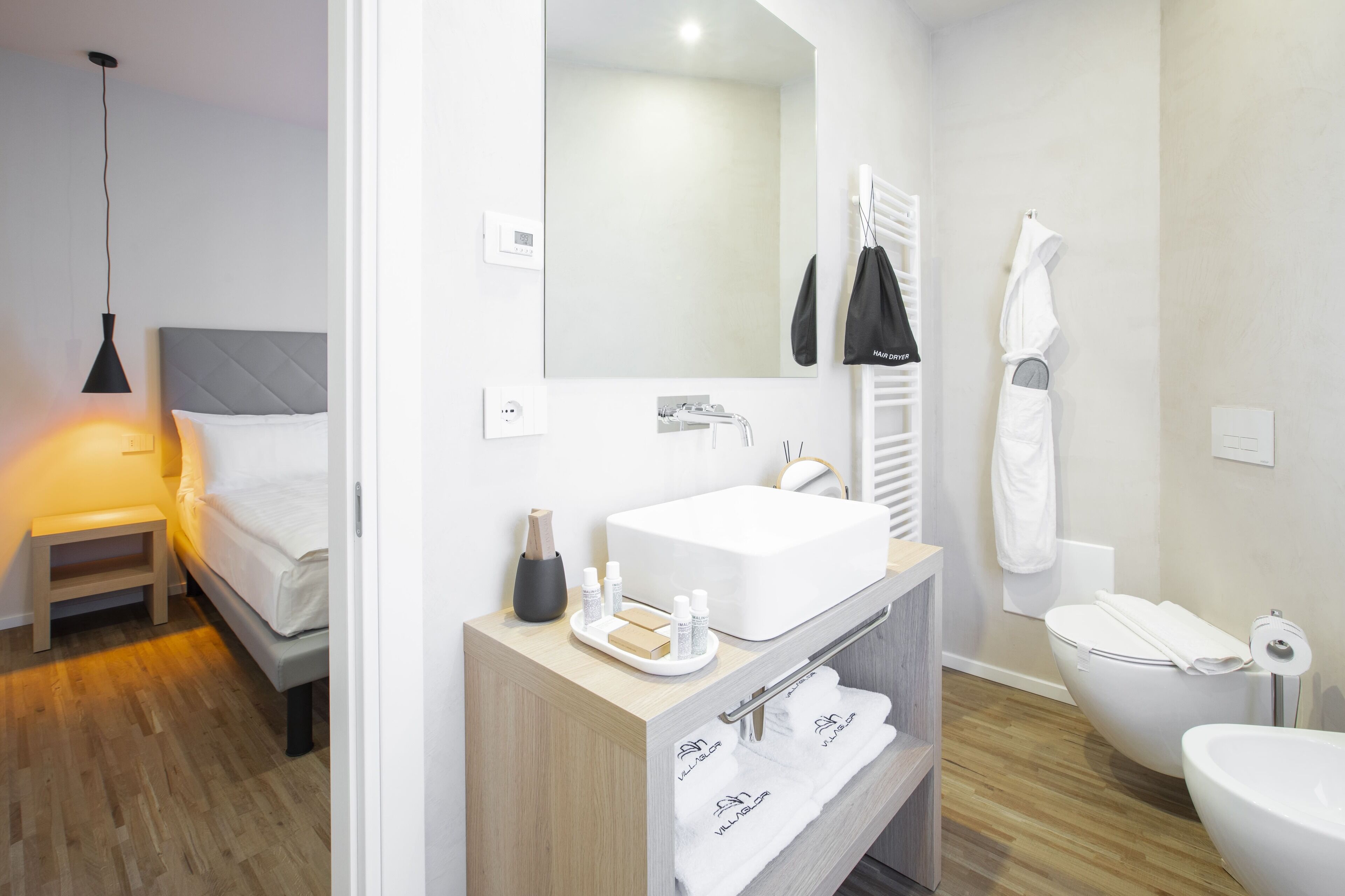 Un bagno moderno con lavandino, specchio e WC bianchi. Un mobile bagno in legno con portasciuga e una piccola mensola.