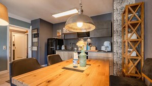 Un coin repas avec une table en bois, des chaises, un luminaire suspendu et des appareils de cuisine en arrière-plan.