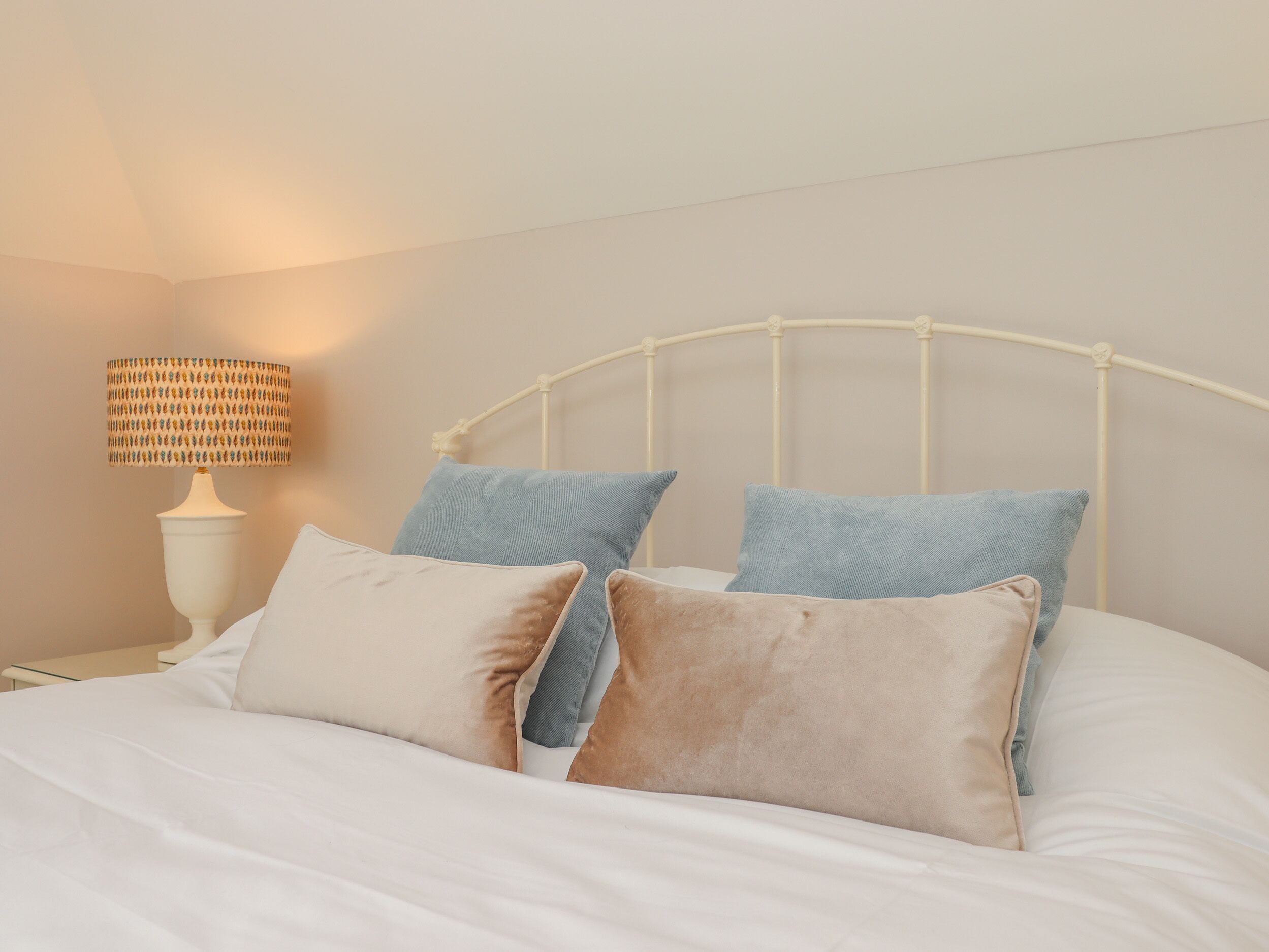 A neatly made bed with white linens and multiple pillows, a white metal headboard, and a lamp with a patterned shade on a bedside table.