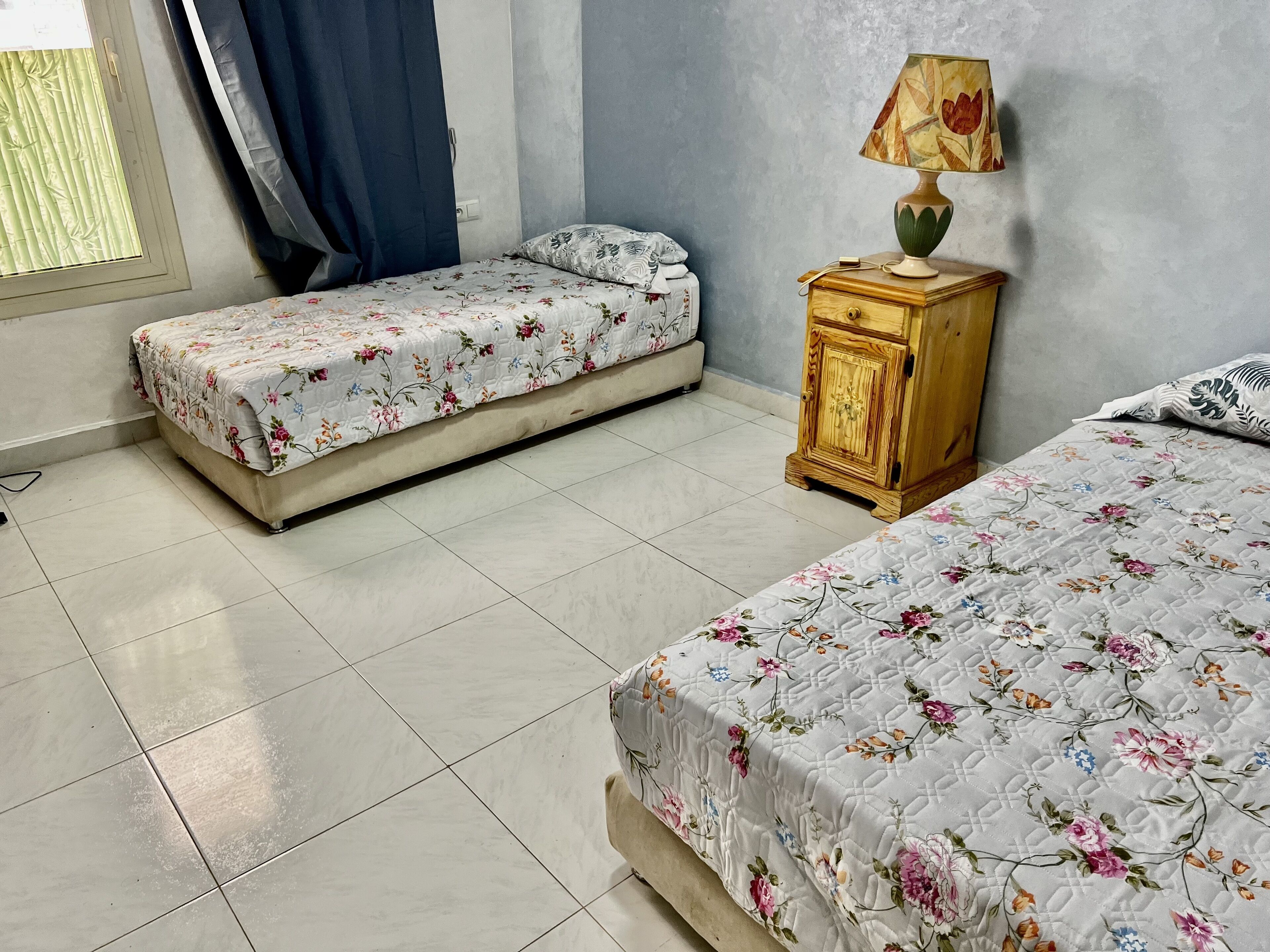 Un dormitorio con dos camas, una mesita de noche de madera, una lámpara y una ventana con cortinas.