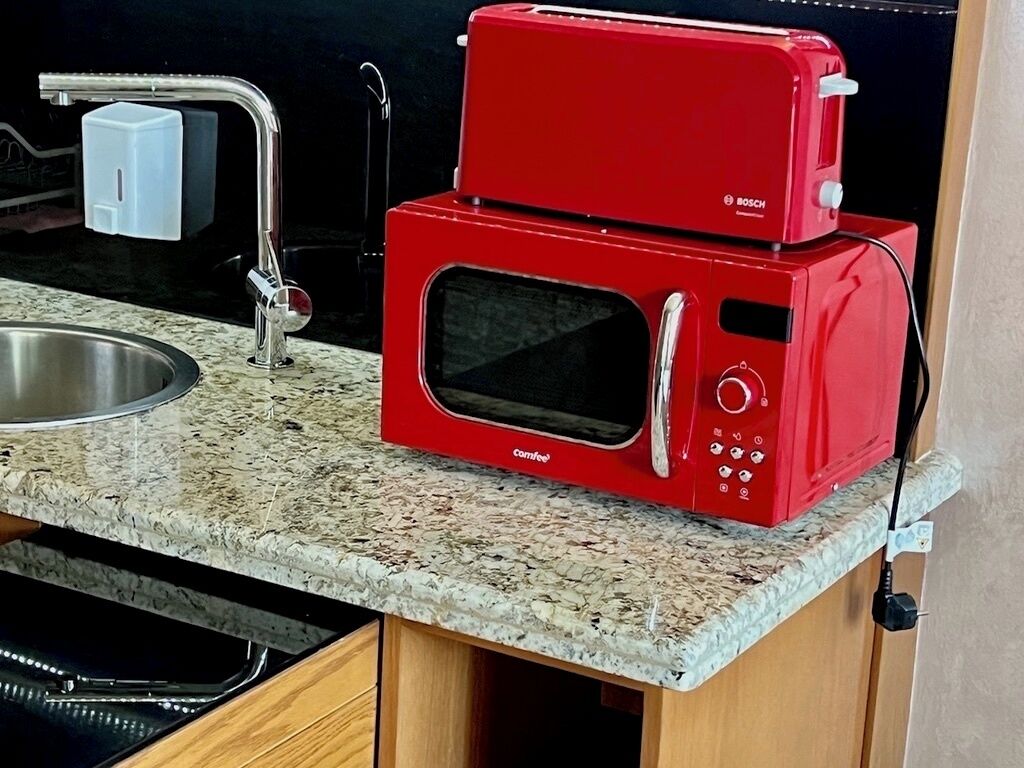 A red microwave oven with a digital display and control panel, placed on a granite countertop next to a stainless steel sink.