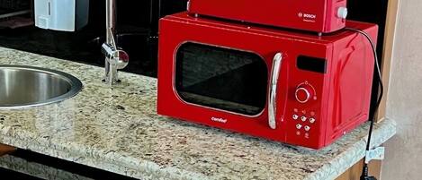 A red microwave oven with a digital display and control panel, placed on a granite countertop next to a stainless steel sink.